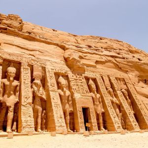 La fachada de un templo antiguo tallado en un acantilado rocoso, con estatuas colosales y jeroglíficos bajo un cielo despejado.