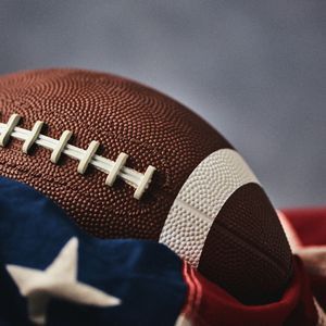 Primo piano di un pallone da football americano marrone con lacci bianchi adagiato su una bandiera americana drappeggiata.
