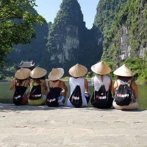 Un groupe WeRoad est assis sur une corniche rocheuse, coiffé de chapeaux coniques et admirant une rivière entourée de montagnes verdoyantes.