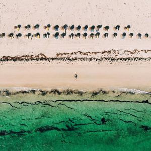 Vue aérienne d'une personne marchant sur une plage de sable blanc, à côté d'une eau turquoise et de rangées de parasols en paille.