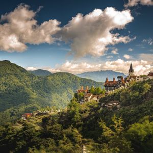 A village with Tudor-style buildings is perched on a lush green mountainside under a blue sky with white clouds.
