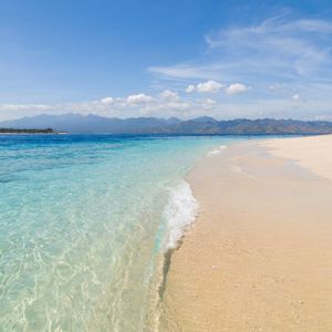 L'eau turquoise et limpide clapote sur une plage de sable blanc avec des montagnes au loin sous un ciel bleu.
