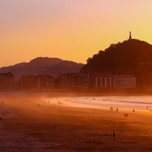 A wide beach filled with people enjoying the orange glow of sunset, with city buildings and a large hill in the background.