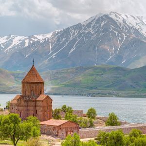 Una chiesa in pietra con tetto conico sorge su una penisola verde accanto a un lago, con una grande montagna innevata sullo sfondo.