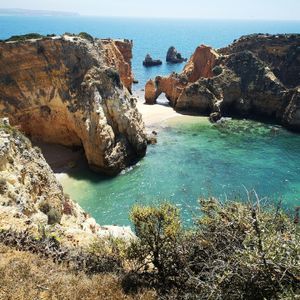 A secluded cove with a natural rock arch is surrounded by golden cliffs and turquoise water, viewed from above on a sunny day.