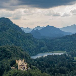 Un castello giallo si erge su una collina boscosa, con un lago e strati di montagne sullo sfondo sotto un cielo nuvoloso.