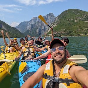 Un viaje en grupo de WeRoad se toma una selfie sonriente mientras hacen kayak en un lago con montañas verdes de fondo.