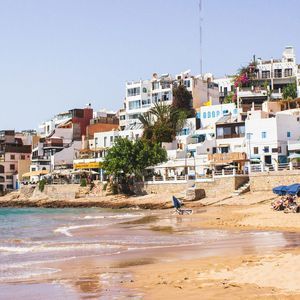 Une ville côtière aux bâtiments blancs surplombe une plage de sable où les gens se détendent sous des parasols bleus.
