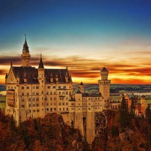 A large castle with multiple towers sits on a forested cliff overlooking a valley with lakes under a colorful sunset sky.
