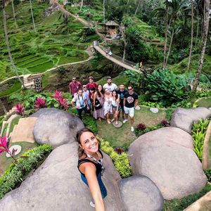 Una mujer se toma una selfie desde un ángulo alto con un grupo de WeRoad posando en una ladera verde aterrazada con grandes rocas.