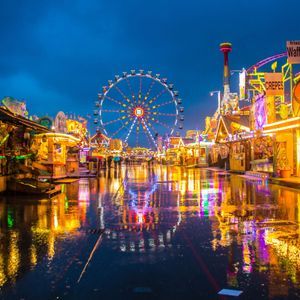 Un parque de atracciones iluminado al anochecer con una noria y puestos de comida, sus luces de colores reflejándose en la calle mojada.