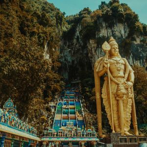 Eine große goldene Statue einer Gottheit steht neben einer langen, farbenfrohen Treppe, die einen bewaldeten Felsen hinaufführt.