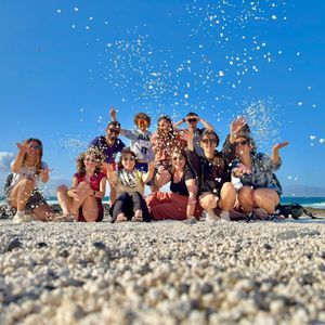 Un grupo de WeRoad lanza guijarros blancos al aire posando para una foto en una playa soleada.