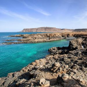 Une côte déchiquetée et rocheuse encadre une étroite crique aux eaux turquoise éclatantes, sous un ciel bleu parsemé de légers nuages.