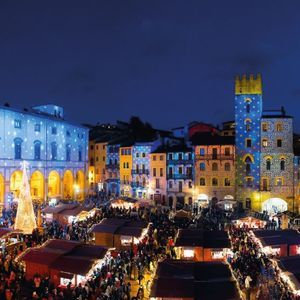 Vista aerea di un affollato mercatino di Natale in una piazza cittadina di notte, con edifici illuminati da proiezioni di luci colorate.