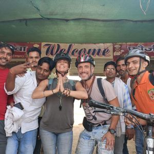 Una persona con un casco è seduta su una mountain bike su un sentiero sterrato, vicino a un ponte di cemento, con vista su un fiume e colline verdi.
