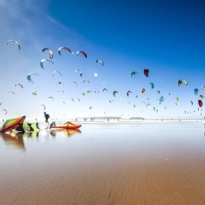 Numerosi aquiloni colorati da kitesurf riempiono un cielo azzurro limpido sopra una spiaggia bagnata e riflettente con persone.