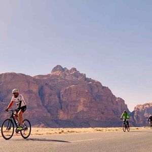 Un viaggio di gruppo WeRoad con persone in bicicletta su una strada asfaltata attraverso un paesaggio desertico con grandi formazioni rocciose sullo sfondo.