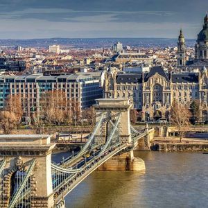 Un grand pont suspendu enjambe une rivière devant un paysage urbain dense, comprenant des bâtiments historiques et une cathédrale à dôme.