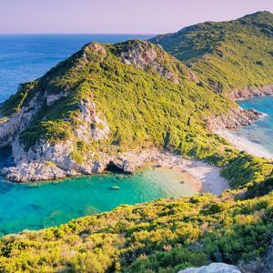 A lush green, rocky headland forms a small cove with a sandy beach and clear, turquoise water on a sunny day.