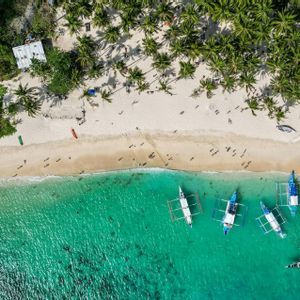 Eine Luftaufnahme eines tropischen Strandes mit weißem Sand, Palmen und Booten, die im türkisfarbenen Wasser ankern.