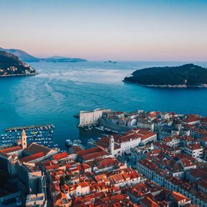 Una vista aerea di una storica città costiera con tetti in tegole rosse, un porto con barche e un'isola nel mare blu all'alba.