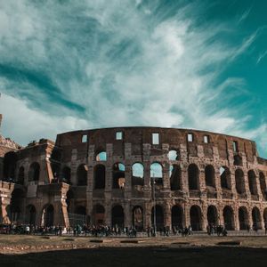 Un grande e antico anfiteatro in pietra si erge sotto un cielo turchese pieno di nuvole leggere, con una folla di persone radunata davanti.