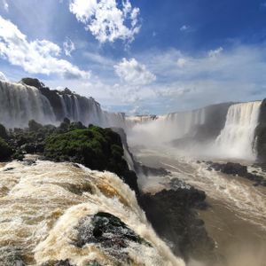 Inmensas cascadas caen sobre acantilados rocosos y exuberantes hacia un cañón neblinoso bajo un cielo azul con nubes blancas.