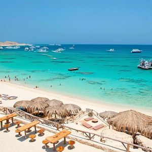 Una spiaggia di sabbia bianca con ombrelloni di paglia si affaccia su una baia dalle acque limpide e turchesi, con numerose barche ancorate al largo.