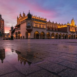 Una vista en ángulo bajo de edificios históricos iluminados en una plaza empedrada al atardecer, reflejados en un charco en el pavimento mojado.