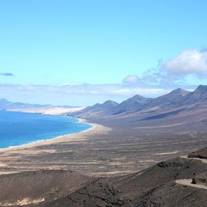 Vista aérea de una costa volcánica con una larga playa de arena que se une al océano azul, y una carretera sinuosa en primer plano.