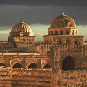 Ancient stone city rooftops and domes illuminated by golden sunlight under a dark, stormy sky.