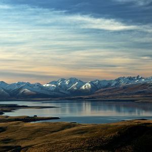 A calm lake reflects a range of snow-capped mountains under a colorful sky, with grassy hills in the foreground.