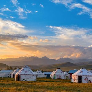 A camp of traditional white yurts in a grassy field with distant mountains under a colorful sunset sky.