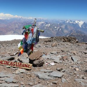 A wooden sign for Aconcagua rests on a rocky summit next to a post with colorful flags, with snowy mountain peaks in the distance.