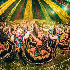Bailarines con elaborados y coloridos disfraces danzan en círculo en un festival, con confeti en el suelo y serpentinas en el aire.