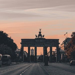 La Puerta de Brandeburgo se alza silueteada al final de una calle ancha contra un cielo anaranjado al atardecer, con la Torre de Televisión de Berlín al fondo.