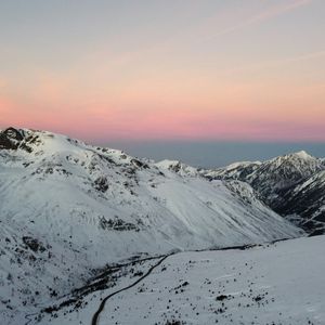 Una vista aérea de montañas cubiertas de nieve con una carretera sinuosa a través de un valle bajo un cielo rosa y azul al anochecer.