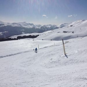 Uno sciatore in giacca blu scende su un vasto pendio innevato, con una catena montuosa in lontananza sotto un cielo soleggiato.