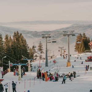 Una frequentata località sciistica con persone sulle piste innevate, seggiovie in alto e uno sfondo di foreste di pini e montagne nebbiose.