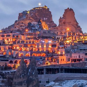 Une vue panoramique d'une ville enneigée construite à flanc de colline rocheuse, illuminée par des lumières chaleureuses au crépuscule.