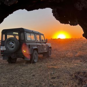 Un SUV scuro è parcheggiato in un campo erboso con il sole che tramonta all'orizzonte, incorniciato da un ramo d'albero in silhouette.