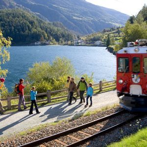 Un treno rosso corre su un binario accanto a un lago di montagna, mentre un gruppo di persone su un sentiero lo saluta.