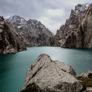 Un lac turquoise serpente à travers un canyon de montagne accidenté avec des sommets enneigés sous un ciel nuageux.
