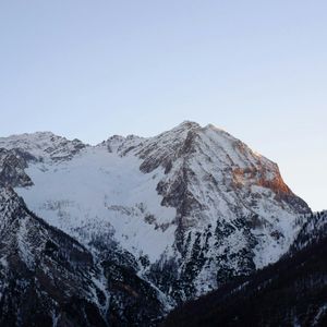 Un sommet enneigé est illuminé par la lumière orange du lever du soleil sous un ciel clair, avec des pentes boisées sombres en contrebas.