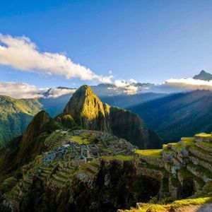A panoramic view of ancient stone ruins and terraces on a green mountainside with a prominent peak lit by sunlight.
