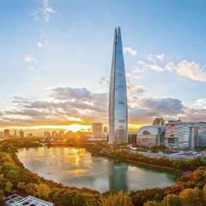 Un alto grattacielo di vetro si erge su uno skyline cittadino accanto a un lago circondato da alberi autunnali al tramonto.