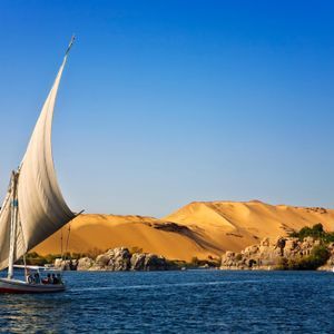 Una tradizionale feluca con persone a bordo naviga su un fiume blu, con dune di sabbia e sponde rocciose sotto un cielo sereno.