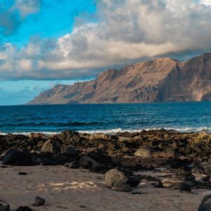 Una costa rocosa con arena y piedras volcánicas se enfrenta a una gran cordillera al otro lado del océano azul bajo un cielo parcialmente nublado.
