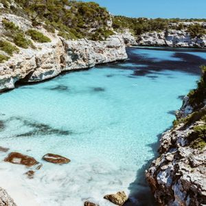 Una vista desde un acantilado alto de una cala apartada con agua turquesa transparente, una pequeña playa de arena blanca y costas rocosas.